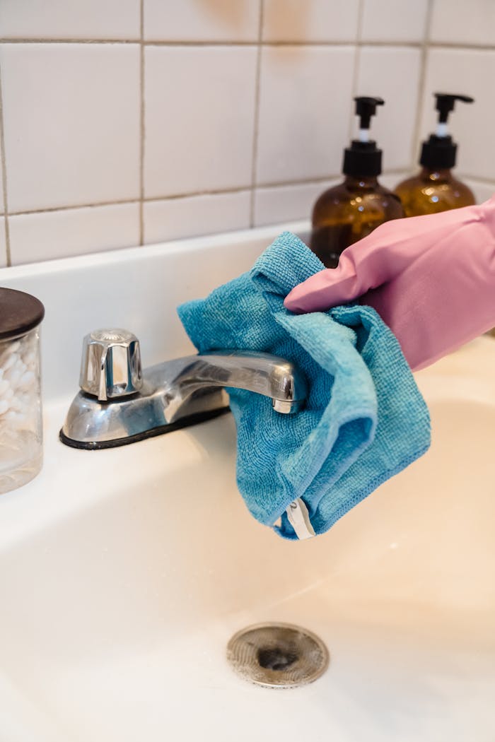 service-01 Close-up of a person sanitizing a bathroom faucet using a blue cloth and latex gloves.
